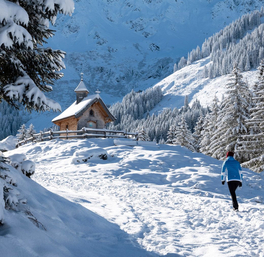 Winter hiker walking on a snowy path near a small mountain chapel