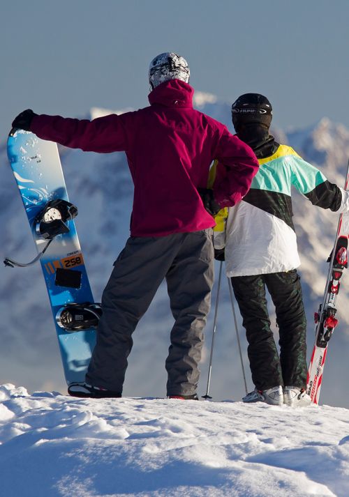 Skiers and snowboarders standing on a snowy mountain peak overlooking the Alps
